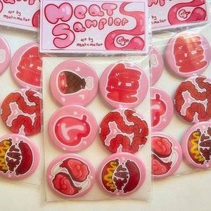 May include: A set of 12 pink and red button pins with various meat-themed illustrations. The pins feature images of a turkey leg, intestines, hot dogs, and other meat-related designs. The pins are packaged in a clear plastic bag with a pink and white header card that reads "Meat Sampler" and "art by meatnmullet".