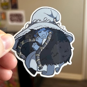 May include: A sticker featuring a blue-skinned character with a large hat and dark hair. The character is depicted in a seated position, wrapped in a striped scarf. The sticker has a white border and is held by a hand.