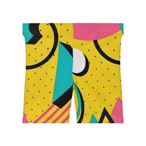 May include: Biker shorts featuring a vibrant yellow base with a repeating pattern of black polka dots, teal geometric shapes, pink diagonal stripes, and solid pink and white circles. The shorts have a high waistband and a fitted silhouette.