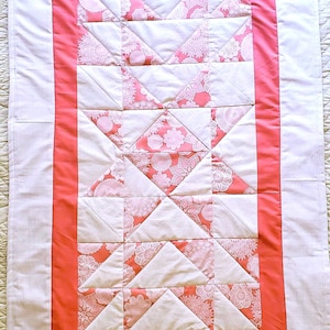 May include: A rectangular, quilted table runner with a central design of white triangles and floral patterned triangles in shades of pink and coral. The runner is bordered by a coral band and white fabric.