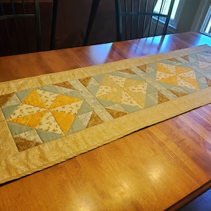 May include: A beige table runner with a quilted design featuring a repeating pattern of blue, brown, and yellow stars. The runner is on a wooden table.
