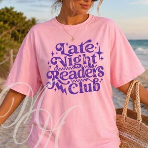 May include: A light pink short-sleeve t-shirt featuring the purple text Late Night Readers Club in a stylized, wavy font. The design includes small star and lightning bolt graphics, along with a checkered pattern element integrated into the text.