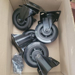 May include: A set of black swivel caster wheels with mounting plates, inside a cardboard box. The wheels have a black rubber tread and are designed for furniture or equipment. A small bag of hardware is also visible.