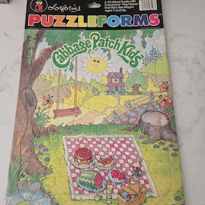 Könnte beinhalten: Ein 30-teiliges Colorforms Puzzleforms Cabbage Patch Kids Puzzle mit einer Picknickszene, einer rot-weiß karierten Decke, einem Kuchen, einer Wassermelone, einem Hotdog und einem Korb. Die Illustration zeigt ein Haus, eine Schaukel und eine Sonne.