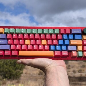 May include: A colorful keyboard with a red frame and rainbow colored keys. The keyboard has a number row, a function row, and a row of keys with arrows and other symbols. The keyboard is being held in a person's hand.