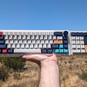 May include: A white mechanical keyboard with a blue, teal, and orange color scheme. The keyboard has a number pad on the right side. The keyboard is being held in a hand against a background of a grassy field and a blue sky.