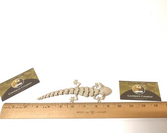 May include: A beige plastic gecko skeleton model, approximately 6 inches long, is lying on a white surface. A ruler is in the foreground, and two business cards are in the background. The business cards have the text "Gavin Schatz Content Creator" on them.