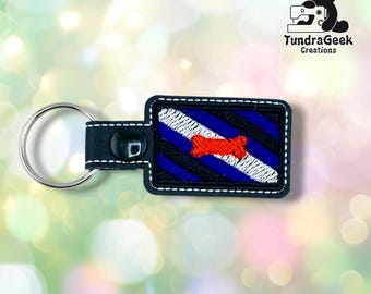 May include: A rectangular black leather keychain featuring a silver metal ring and white stitching. The center displays an embroidered pattern of diagonal blue and white stripes with a small, bright orange bone shape centered on the design.