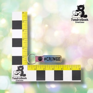 A pink and blue ombre keychain with "#CRINGE" text displayed on a yellow measuring tape next to TundraGeek Creations branding.