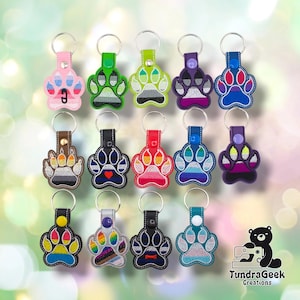 May include: A collection of fourteen embroidered paw print keychains, each featuring a unique color pattern and a metal split ring. The keychains are arranged in three rows and include a small logo of a bear with a sewing machine in the bottom right.