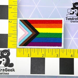 Puede incluir: Un parche blanco con una bandera arco&iacute;ris del orgullo con un tri&aacute;ngulo negro, marr&oacute;n, azul claro, rosa y blanco en la esquina superior izquierda. El parche mide 5 cm de ancho y 7,5 cm de largo. TundraGeek Creations est&aacute; impreso en texto negro sobre fondo blanco.