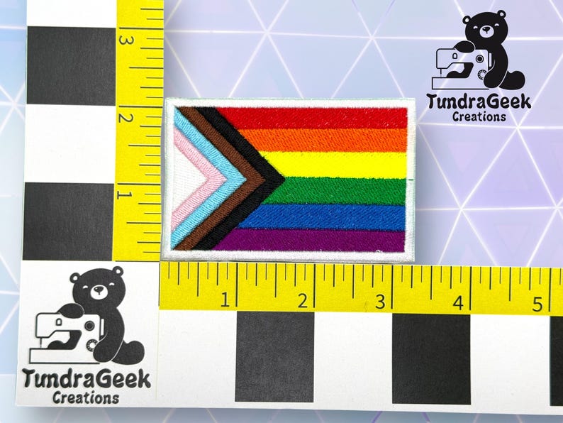 Puede incluir: Un parche blanco con una bandera arco&iacute;ris del orgullo con un tri&aacute;ngulo negro, marr&oacute;n, azul claro, rosa y blanco en la esquina superior izquierda. El parche mide 5 cm de ancho y 7,5 cm de largo. TundraGeek Creations est&aacute; impreso en texto negro sobre fondo blanco.