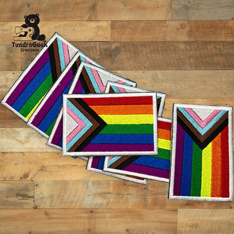 Puede incluir: Un mont&oacute;n de parches bordados con la bandera del Orgullo Progresivo, una variaci&oacute;n de la bandera del arco&iacute;ris que incluye rayas negras, marrones, azul claro y rosas para representar a los grupos marginados. Los parches tienen un borde blanco y un aspecto texturizado y bordado.