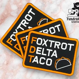 Puede incluir: Tres parches bordados negros con bordes naranjas. Los parches presentan el texto "FOXTROT" y "DELTA TACO" en blanco, junto con un gráfico de taco. Los parches son de TundraGeek Creations.