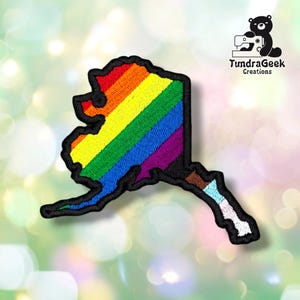 Puede incluir: Un parche bordado con la forma del mapa de Alaska, con rayas diagonales en rojo, naranja, amarillo, verde, azul y morado, con un borde negro. La sección inferior incluye segmentos en marrón, azul claro y blanco. Texto: TundraGeek Creations.