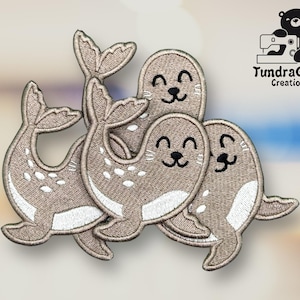 Puede incluir: Parches bordados con un grupo de focas sonrientes en tonos beige y blanco. Los parches tienen un diseño cosido detallado, con el texto "TundraGeek Creations" y un logotipo de oso en la esquina superior derecha.