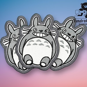 May include: Three gray embroidered iron-on patches of the character Totoro from the movie My Neighbor Totoro. Each patch features a different expression on Totoro's face.