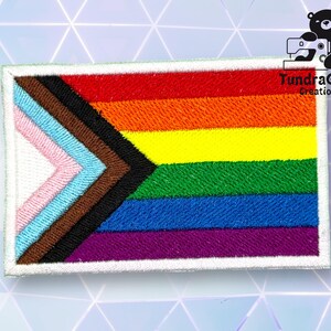 Puede incluir: Un parche blanco con un borde blanco presenta una bandera del orgullo arco&iacute;ris con un dise&ntilde;o de chevron en el centro. El chevron est&aacute; formado por seis colores: rosa, azul claro, marr&oacute;n, negro, azul oscuro y morado. El texto "TundraGeek Creations" est&aacute; en la esquina superior derecha del parche.