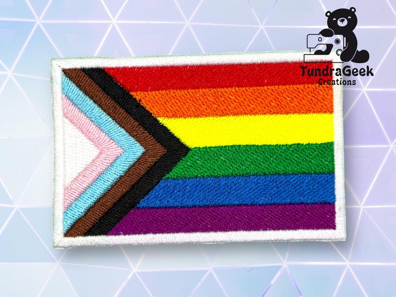 Puede incluir: Un parche blanco con un borde blanco presenta una bandera del orgullo arco&iacute;ris con un dise&ntilde;o de chevron en el centro. El chevron est&aacute; formado por seis colores: rosa, azul claro, marr&oacute;n, negro, azul oscuro y morado. El texto "TundraGeek Creations" est&aacute; en la esquina superior derecha del parche.