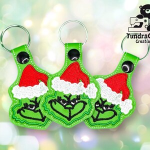 May include: Three green keychains featuring the Grinch's face wearing a red Santa hat with white trim. The keychains have silver key rings and black accents. The brand logo "TundraGeek Creations" is visible in the upper right corner.