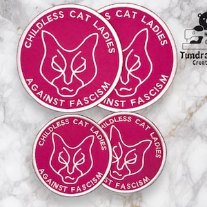 Puede incluir: Cuatro parches bordados termoadhesivos de color rosa y blanco con un diseño de gato y el texto "Childless Cat Ladies Against Fascism".