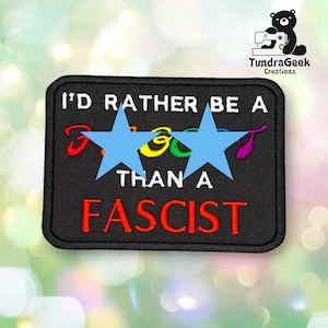 May include: A rectangular black embroidered patch with white stitched text that reads I'D RATHER BE A THAN A FASCIST. The word between the stars is obscured by two light blue star-shaped overlays. The patch has a black stitched border.
