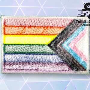 Puede incluir: Una bandera del orgullo arco&iacute;ris con un chevron negro en el centro. La bandera est&aacute; hecha de tela y tiene un borde blanco.