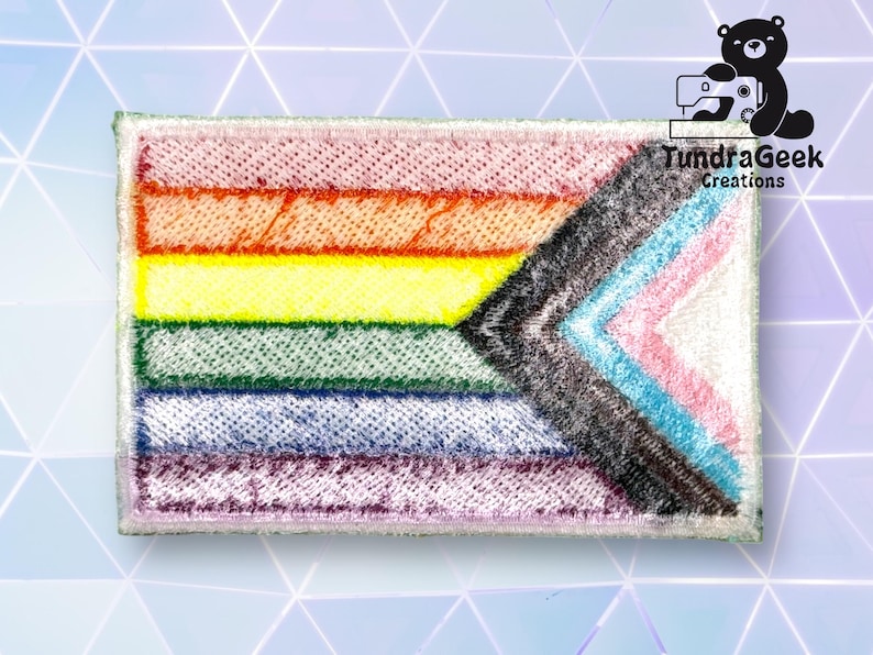 Puede incluir: Una bandera del orgullo arco&iacute;ris con un chevron negro en el centro. La bandera est&aacute; hecha de tela y tiene un borde blanco.