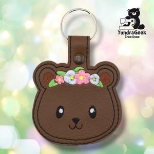 以下が含まれることがあります： 花冠をつけた茶色のクマの形をしたキーホルダー。キーホルダーは、シルバーのキーリングと茶色の革ストラップを備えたステッチデザインです。クマは刺繍のディテールが施されたかわいい顔をしています。「TundraGeek Creations」の文字が見えます。