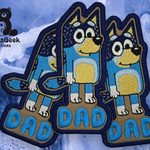 May include: Embroidered patches depicting a cartoon dog in blue, yellow, and white. Each patch has the word "DAD" in blue embroidery on a brown base. The patches are stacked, with the TundraGeek Creations logo in the upper left corner.