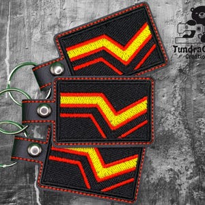 May include: A black leather keychain with three red and yellow embroidered patches. The patches feature a chevron design with a red outline and yellow stripes.