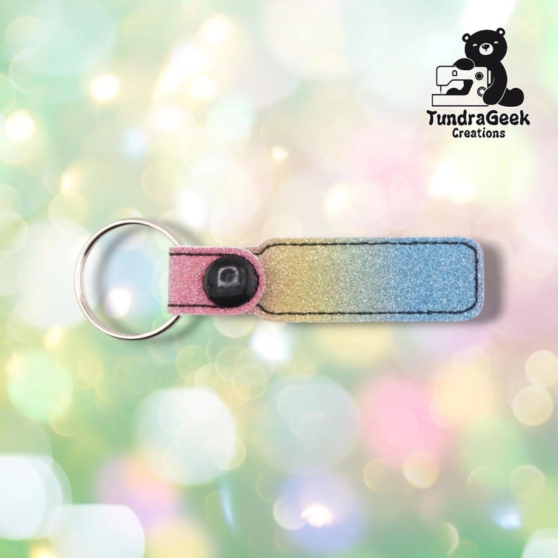 A glittery keychain with pink, gold, and blue gradient ombre finish, metal ring, and black button detail on soft pastel bokeh background.
