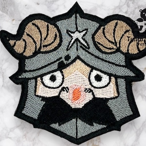 May include: Embroidered patch featuring a cartoon character with horns, a helmet, and a large nose. The character has a light brown, gray, and black color scheme.
