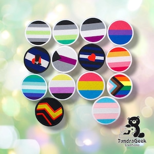 May include: A collection of thirteen circular embroidered patches featuring various pride flag designs, including stripes in colours like pink, blue, yellow, green, purple, and black, with some patches displaying symbols like hearts or geometric shapes.