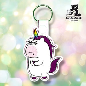 Könnte beinhalten: Ein weißer Einhorn-Schlüsselanhänger mit violetter Mähne und Schweif, einer rosa Schnauze und einem regenbogenfarbenen Horn. Das gestickte Design zeigt einen mürrischen Gesichtsausdruck und ist an einem silbernen Metallring befestigt.