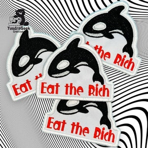 Puede incluir: Parches bordados con un diseño de orca en blanco y negro y el texto rojo "Eat the Rich". Los parches tienen un borde blanco y se muestran sobre un fondo a rayas en blanco y negro.