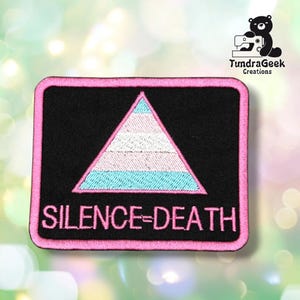 Puede incluir: Un parche rectangular bordado con fondo negro y borde rosa. Presenta un triángulo central con rayas horizontales en azul claro, blanco y rosa, y el texto SILENCE=DEATH en letras mayúsculas de color rosa en la parte inferior.