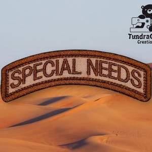 May include: Brown embroidered patch with the text "SPECIAL NEEDS" in a tan color.