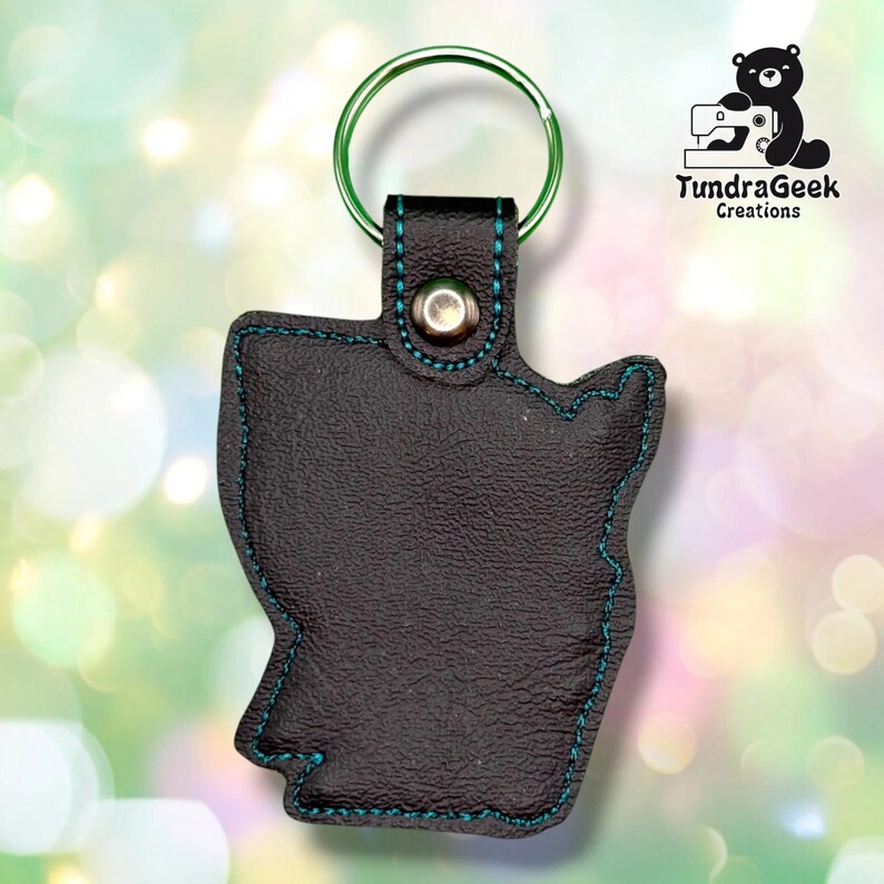 Back of the keychain, showing a plain black vinyl reverse side with teal outline stitching following the hooded figure's silhouette. Green metal split ring and silver snap closure at top.