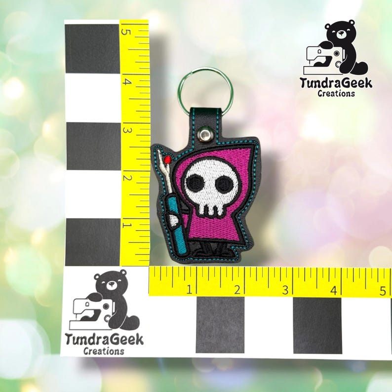 Seam Reaper keychain shown against a black-and-white checkered background with a yellow ruler for scale, measuring approximately 4.25 inches tall by 2.5 inches wide.  ---