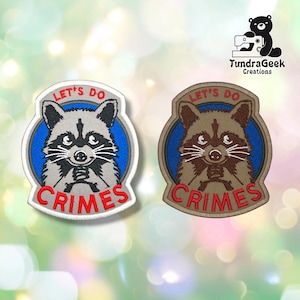 May include: Two embroidered patches featuring a raccoon graphic on a blue circular background. The text LET'S DO CRIMES is stitched in red. One patch has a light gray border and raccoon, while the other has a tan border and brown raccoon.