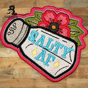 Puede incluir: Salero blanco con una flor rosa y hojas verdes en la parte superior. El salero tiene el texto "SALTY AF" en azul con rayos amarillos a cada lado.