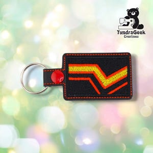 May include: A rectangular black keychain featuring an embroidered design of two parallel yellow and orange zigzag lines. The keychain is finished with red stitching along the edges, a red snap button, and a silver metal split ring for keys.