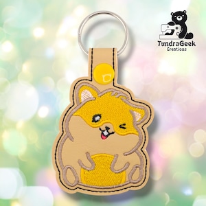 May include: A tan and yellow hamster keychain with a metal ring. The hamster is winking and has a smiling expression. The keychain has a yellow button and the text "TundraGeek Creations".