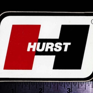 Könnte beinhalten: Ein rechteckiger Aufkleber mit weißem Hintergrund und schwarzem Rand. Der Aufkleber zeigt ein großes, stilisiertes "H", das in rote und schwarze Abschnitte unterteilt ist, mit dem Wort "HURST" in weißer Schrift in der Mitte. Das Warenzeichen befindet sich in der oberen rechten Ecke.
