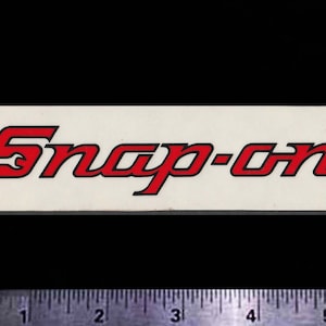 May include: A rectangular sticker with the words "Snap-on" in red, outlined in black, on a white background. The sticker has a black background and a ruler is at the bottom.