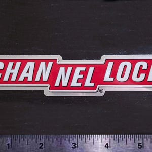 May include: A rectangular sticker with the words "CHANNEL LOCK" in white, bold font. The text is set against a red background with a silver border. The sticker is on a dark surface.
