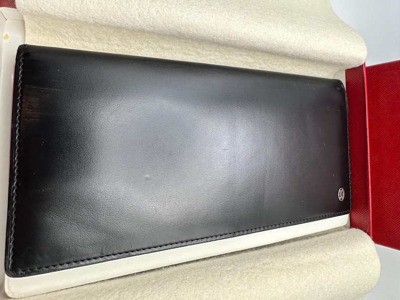 May include: A black leather long wallet with a white accent stitch along the edges. The wallet is displayed in a red box with a beige felt lining.  A subtle logo is visible on the lower right corner. The smooth leather has a slight sheen. This luxury wallet is a sophisticated accessory.
