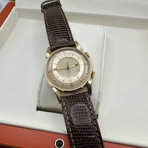 Puede incluir: Un reloj de pulsera de oro con una correa de cuero marr&oacute;n. La esfera del reloj es blanca con marcas negras y un segundero dorado. El reloj est&aacute; en una caja blanca con un forro rojo.