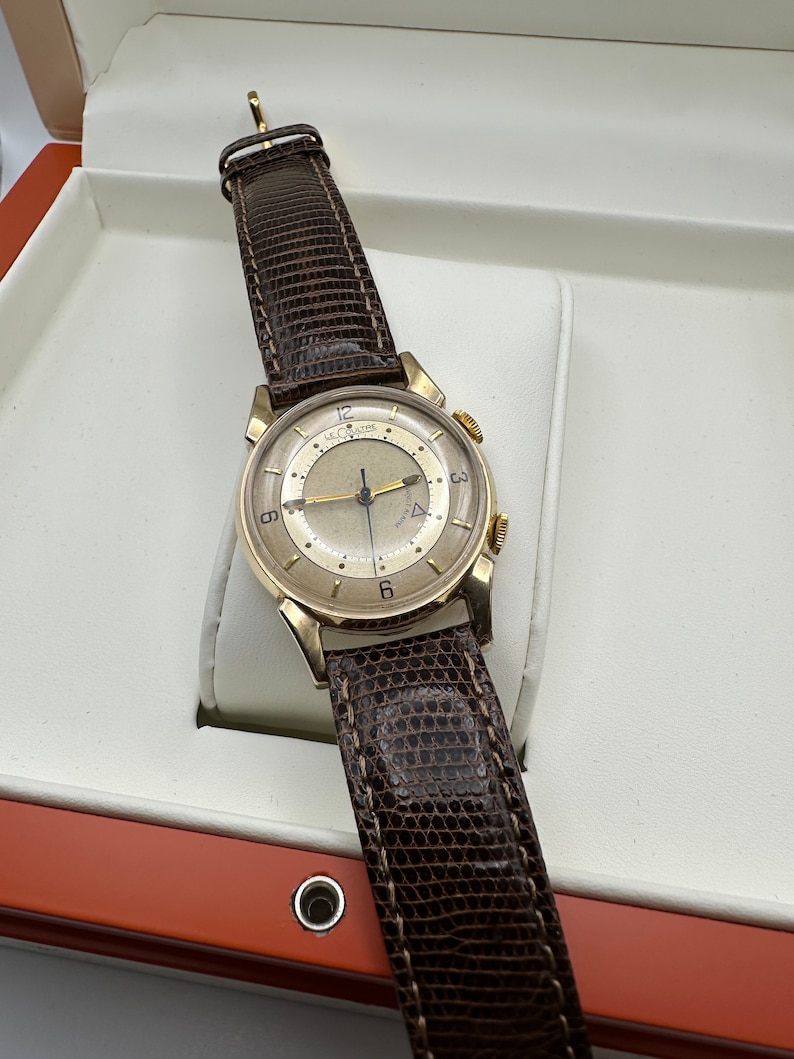 Puede incluir: Un reloj de pulsera de oro con una correa de cuero marr&oacute;n. La esfera del reloj es blanca con marcas negras y un segundero dorado. El reloj est&aacute; en una caja blanca con un forro rojo.